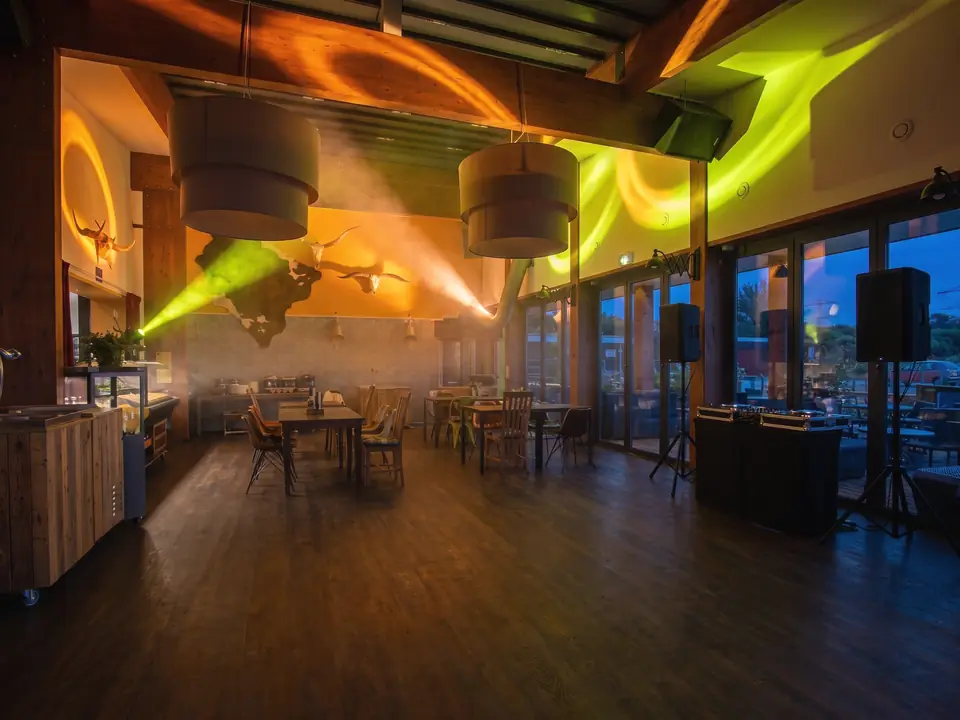 De zaal met houten balken als dansvloer, een DJ-tafel bij de glazen deuren en gekleurd licht door een lichte nevel (AI-beeld)