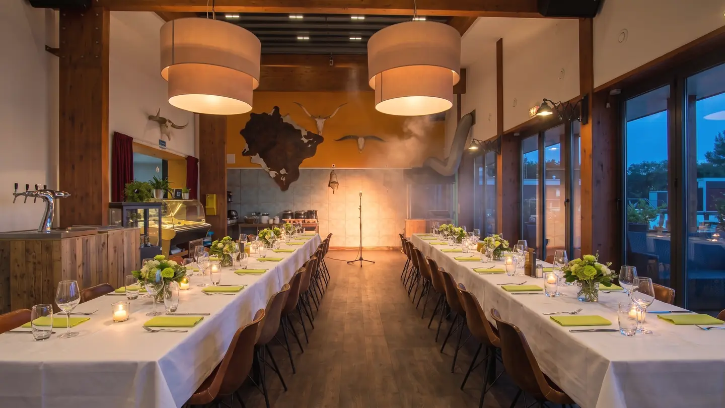 De zaal met houten balken, 's avonds gedekt voor een feest: lange tafels met witte kleden, kaarsen en bloemen, achterin een microfoon in warm licht (AI-beeld)