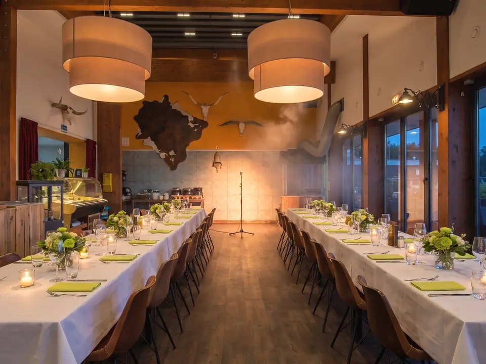 De zaal met houten balken, 's avonds gedekt voor een feest: lange tafels met witte kleden, kaarsen en bloemen, achterin een microfoon in warm licht (AI-beeld)