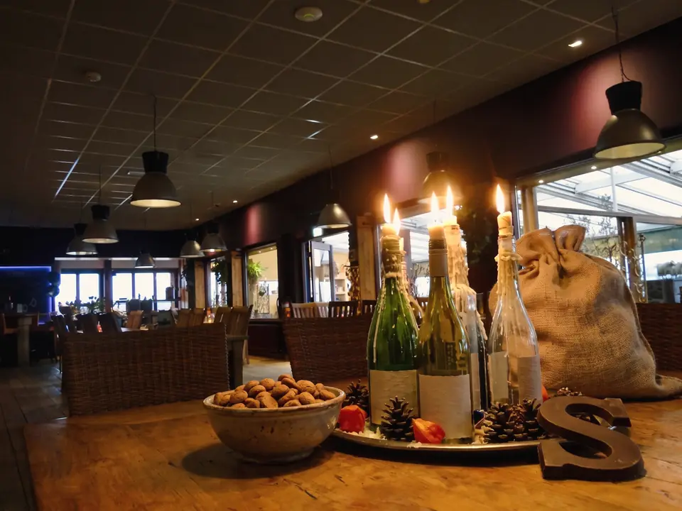 Een tafel met kaarsen in wijnflessen, een schaal pepernoten, een chocoladeletter en een jute zak bij een stoel (AI-beeld)