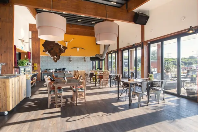 De zaal met houten balken, grote lampen, de oranje wand en glazen deuren naar het terras (AI-beeld)